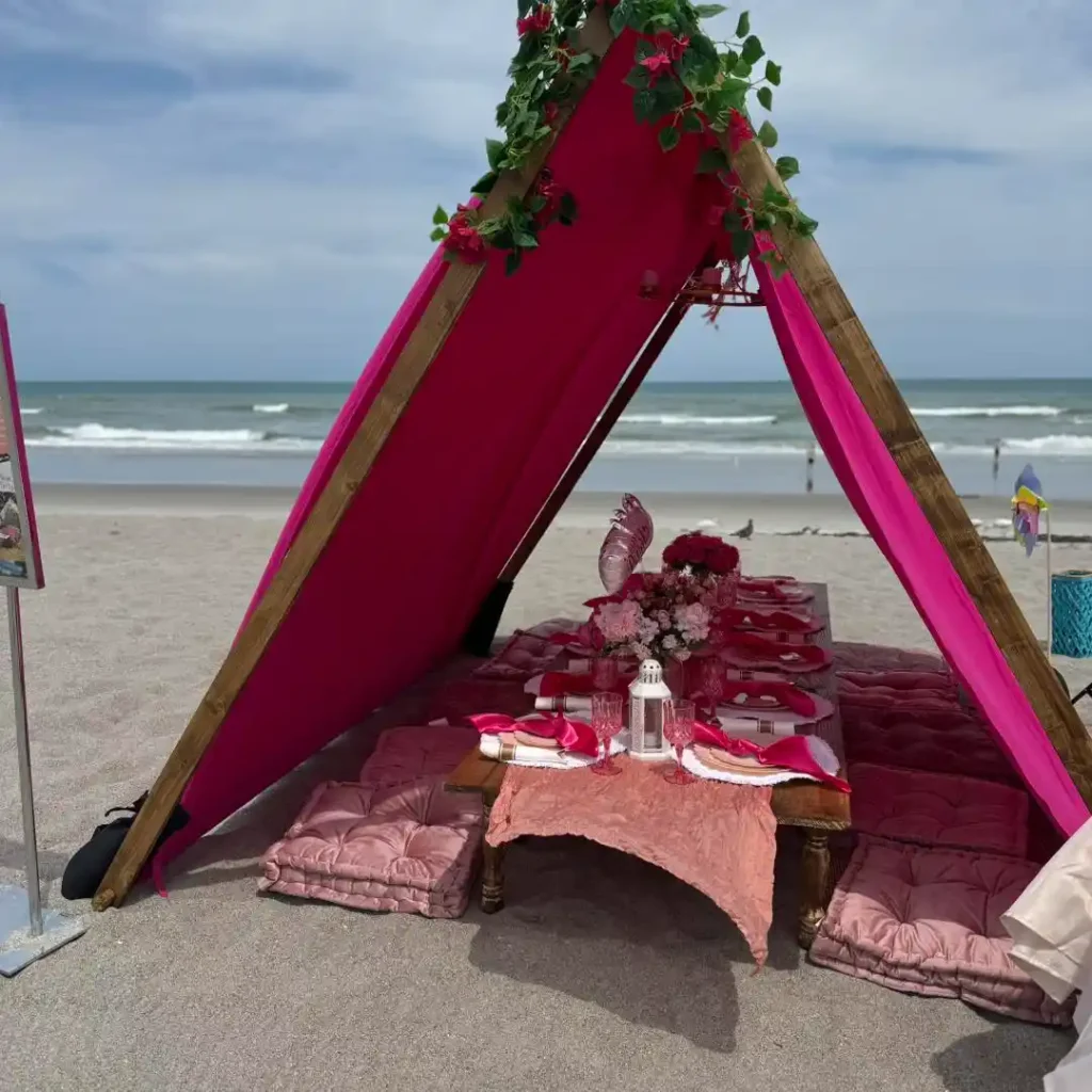 A teepee with pink fabric and flower decorations shelters a low table set for a meal, surrounded by pink cushions, on a sandy beach near the ocean—perfect inspiration from a Central Florida party shop.