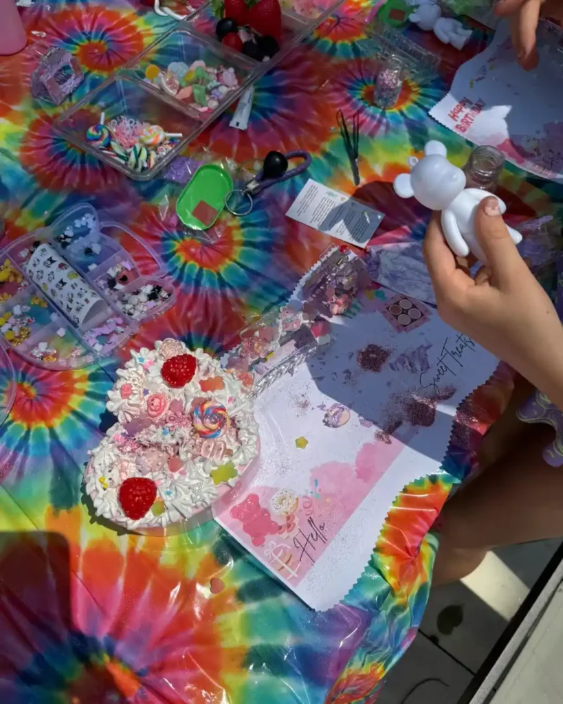 Hands holding a white bear figurine over a tie-dye table filled with craft supplies, decorated pastries, and art materials—a perfect kids activity for creative fun.