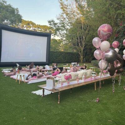 An outdoor movie night in Florida with people lounging on cushions in front of a large screen, surrounded by pink balloons and decorated tables on a grassy lawn.