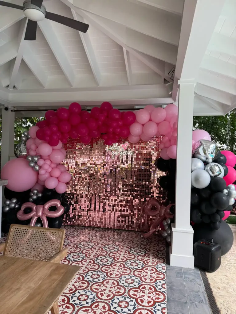 A photo of a party setup, expertly curated by a party planner, featuring a pink sequin backdrop, pink and black balloon arch, and metallic accents under a white gazebo with patterned floor tiles.