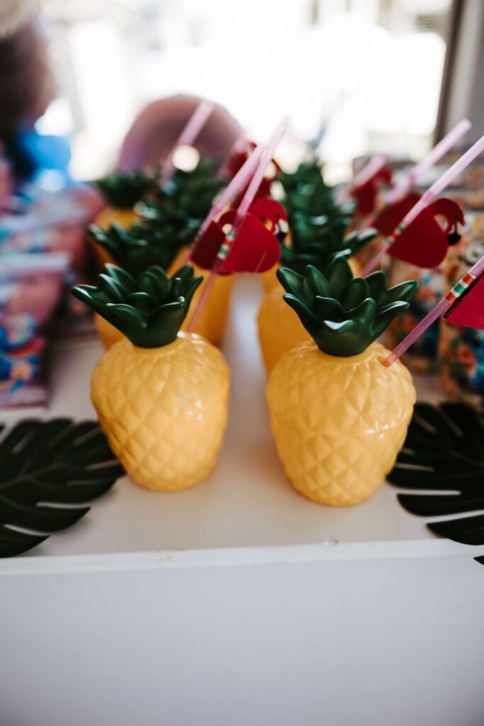 Pineapple-shaped plastic cups with green lids and pink straws are arranged on a table with artificial palm leaves and colorful packages in the background.