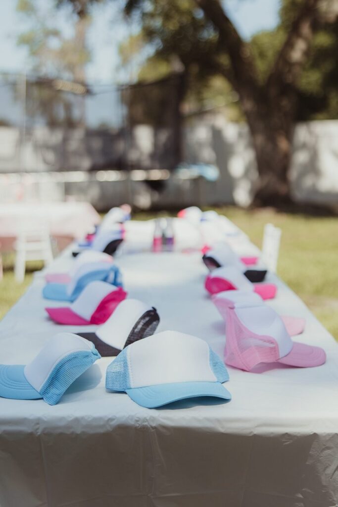 A long table covered with a white tablecloth holds multiple colorful trucker hats, including white, blue, pink, and black, arranged in rows outdoors.