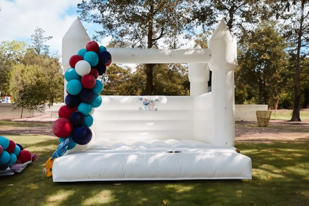 A white inflatable bounce house decorated with colorful balloons stands on grass, surrounded by trees in an outdoor setting.