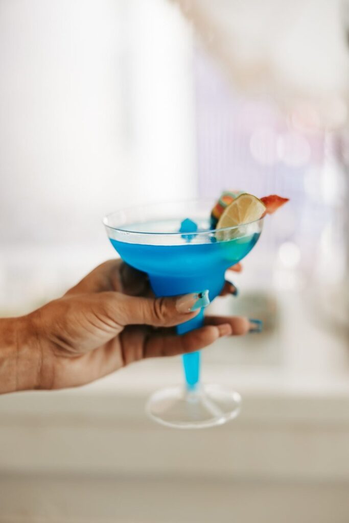 A hand holding a blue cocktail in a glass, garnished with a lime slice and a small piece of fruit.