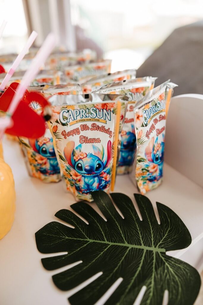Several Capri Sun juice pouches with custom "Happy 5th Birthday Eliana" Stitch-themed labels are arranged behind a decorative green palm leaf.