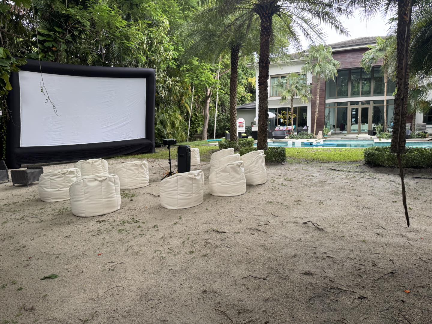 An outdoor movie party setup with white bean bag chairs facing a large screen, surrounded by trees, next to a pool and modern house.