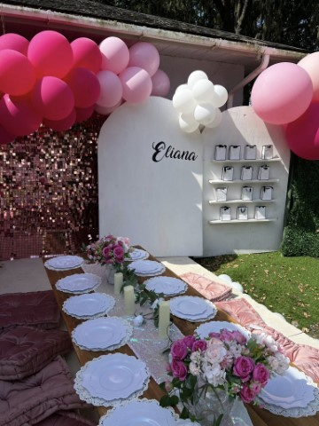 Outdoor party setup inspired by a beach cabana party, featuring pink balloons, floral table decorations, a long table with white plates, and a white backdrop labeled "Eliana" with gift bags on shelves.
