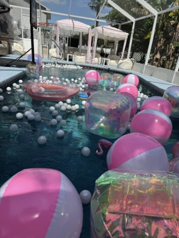A swimming pool filled with pink and white beach balls and inflatable floats sets the perfect scene for a beach cabana party, with stylish white outdoor cabanas and comfortable seating in the background.