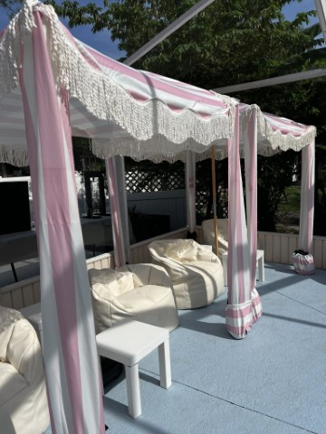 Outdoor seating area with white bean bag chairs, small tables, and pink-and-white striped canopies with fringe, creating a relaxed beach cabana party ambiance under partial shade from nearby trees.